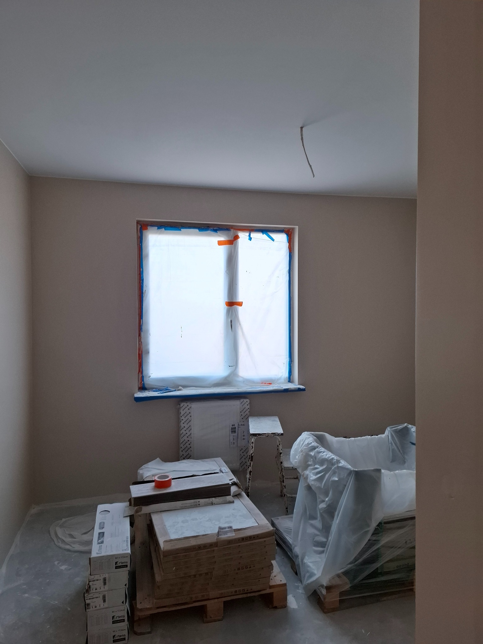 Pokój w trakcie remontu: ściany pomalowane na beżowo, okno zabezpieczone folią, na podłodze kartony z panelami i taśma malarska.