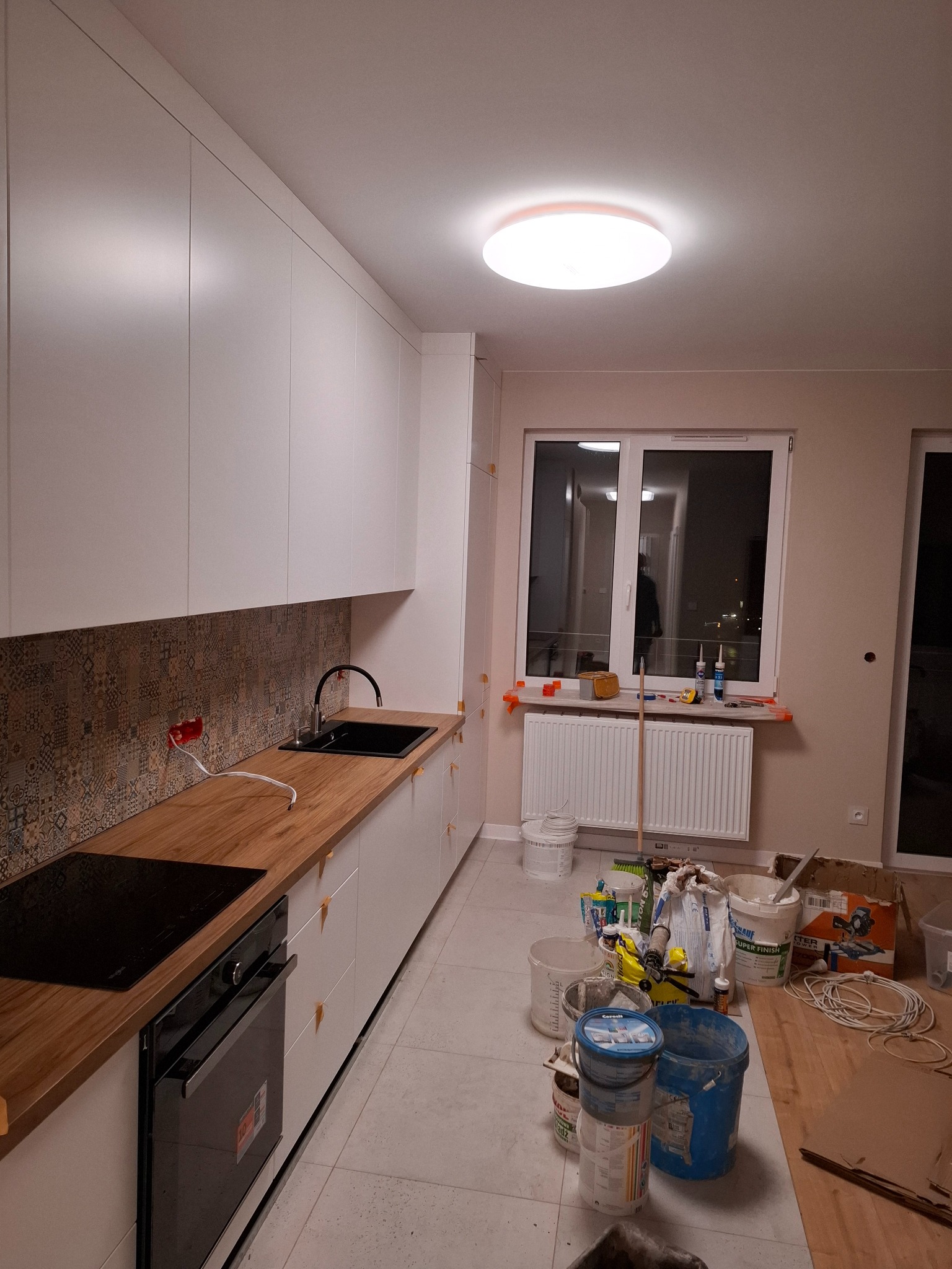 Wnętrze kuchni w trakcie wykańczania: białe szafki z drewnianym blatem, wzorzysta mozaika nad blatem, widoczne narzędzia i materiały budowlane na podłodze, okno z grzejnikiem.