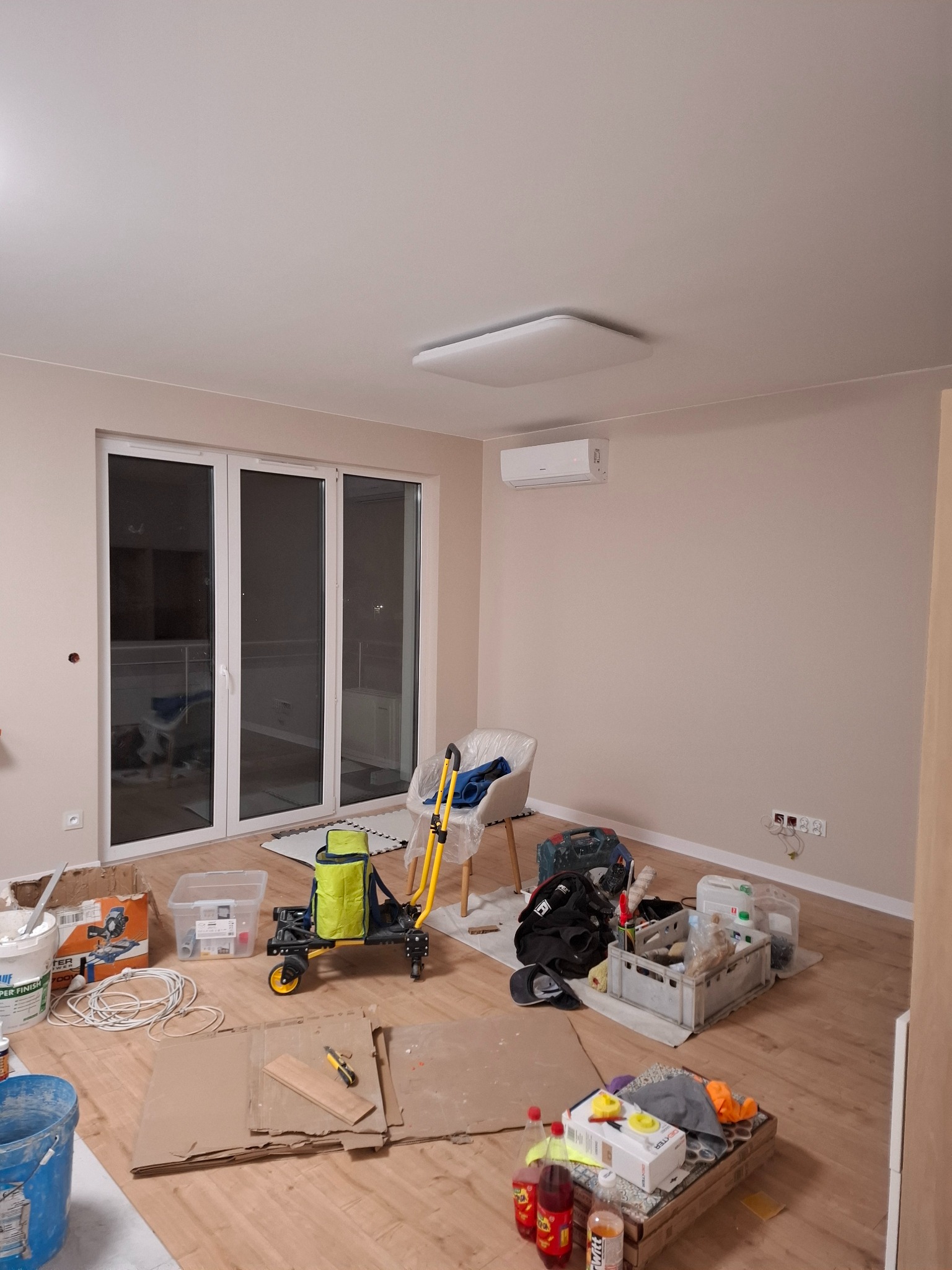 Pokój w trakcie remontu, z widocznymi narzędziami, materiałami budowlanymi, zakryta podłoga kartonem, świeżo pomalowane ściany w odcieniach beżu i zamontowany klimatyzator.