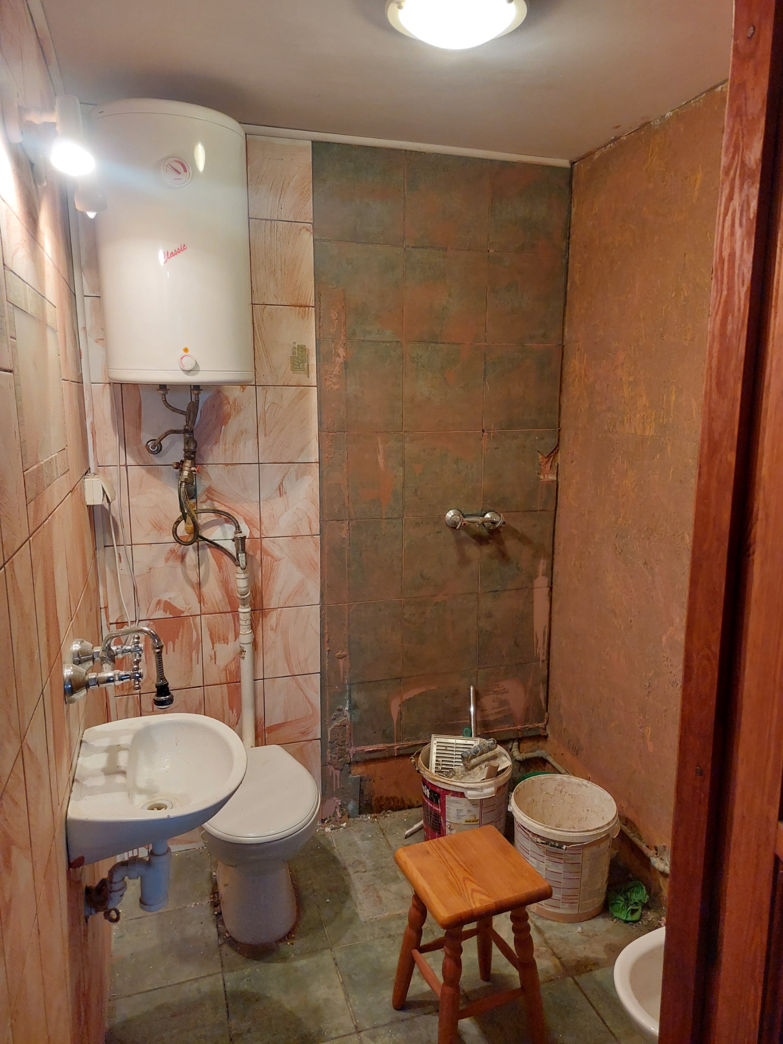 Łazienka w trakcie remontu: ściany częściowo z brązowymi płytkami, częściowo przygotowane do wykończenia, widoczne wiaderka z farbą i stołek.