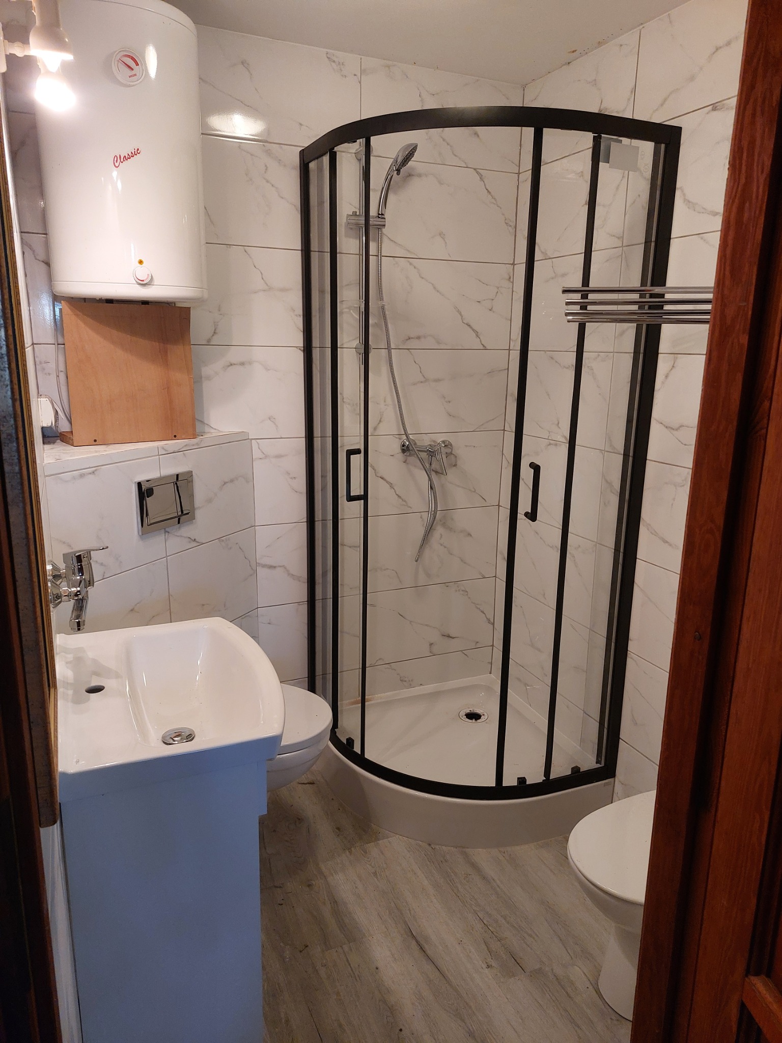 Wykończona łazienka z kabiną prysznicową o czarnych ramach, umywalką z szafką, podwieszaną toaletą i bojlerem Classic na ścianie z marmurkowymi płytkami.