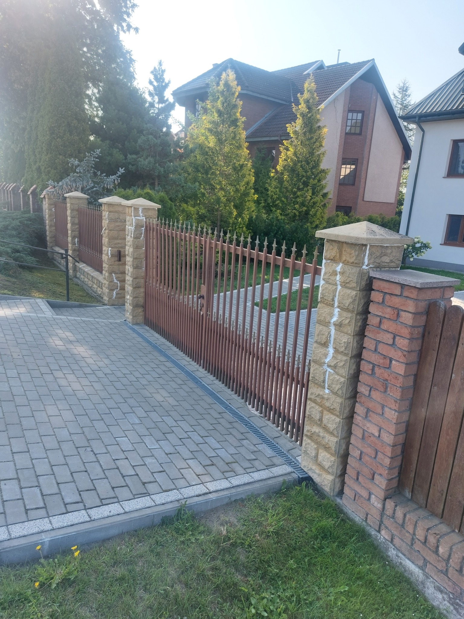Nowoczesne ogrodzenie posesji z metalowymi przęsłami i kamiennymi słupkami, świeżo po fugowaniu, z widoczną kostką brukową na podjeździe i zielenią w tle.