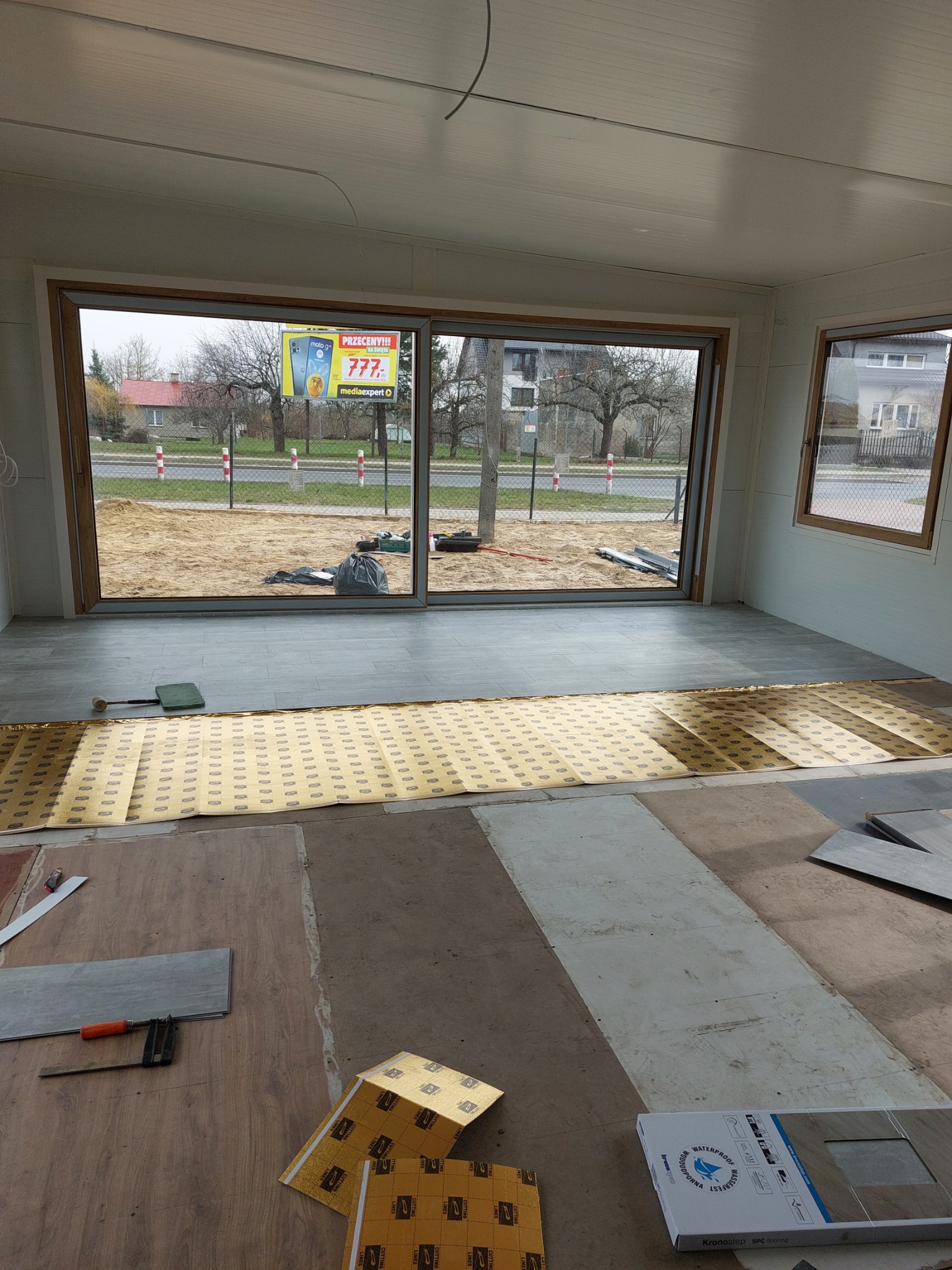 Wnętrze pomieszczenia w trakcie układania podłogi z paneli winylowych; widoczne narzędzia, podkład, częściowo ułożone panele i opakowanie paneli Kronostep SPC flooring.