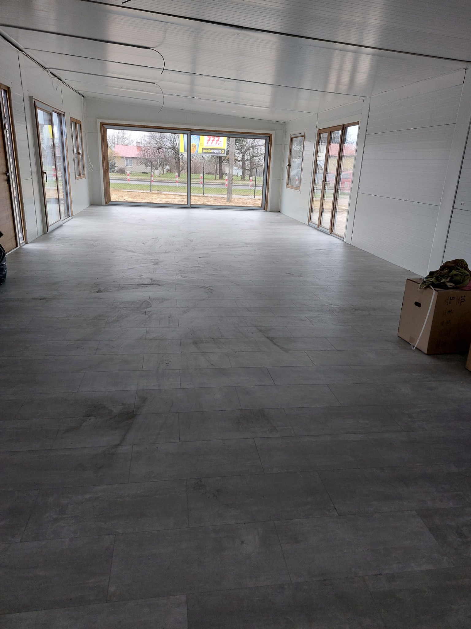 Wnętrze kontenera mieszkalnego z wykończoną podłogą z paneli imitujących kamień, widoczne okna i drzwi balkonowe z drewnianymi ramami, ściany wykończone panelami ściennymi, na podłodze leżą kartony.