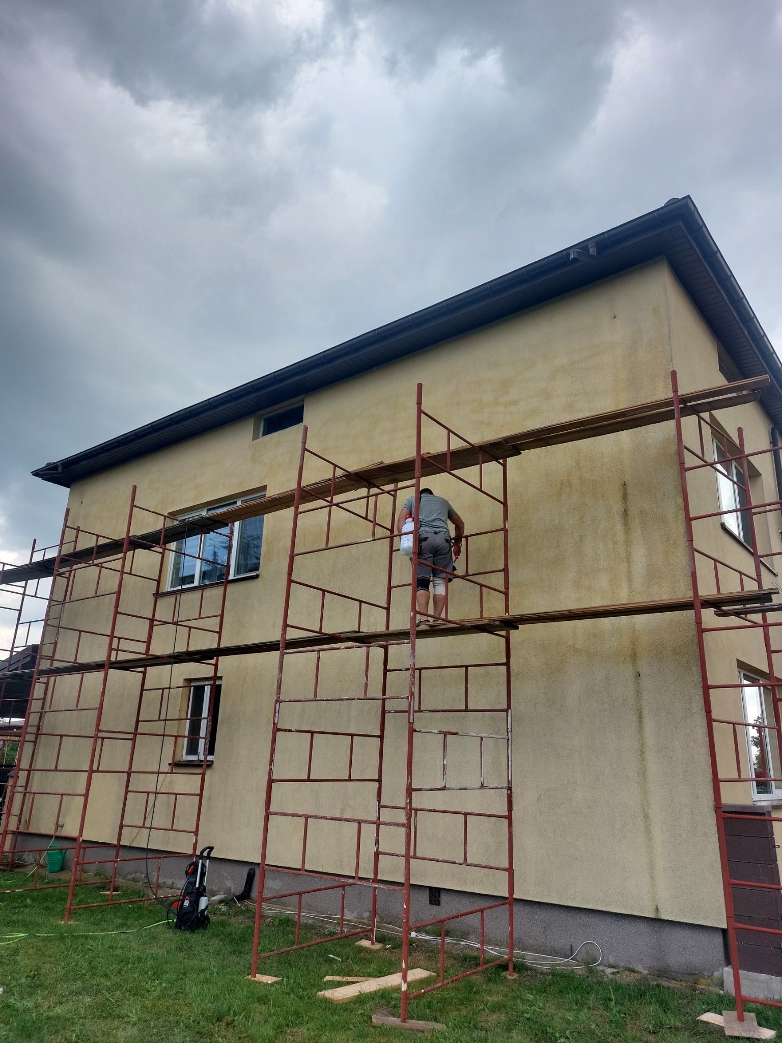 Dwupiętrowy dom w trakcie prac elewacyjnych, widoczny z bliska na rusztowaniu pracownik z pojemnikiem, elewacja w kolorze beżowym, częściowo zabrudzona, pochmurne niebo w tle.