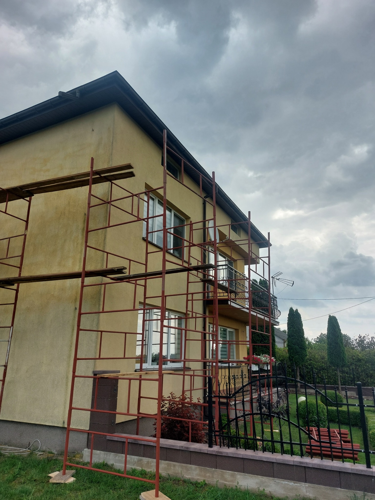 Dwupiętrowy dom w trakcie remontu elewacji, otoczony rusztowaniem, widok z poziomu trawnika na tle pochmurnego nieba.