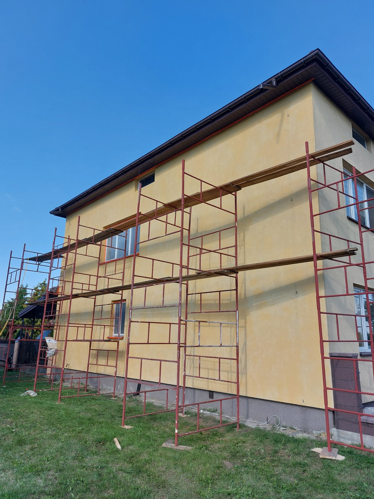 Dwupiętrowy dom w trakcie renowacji elewacji, otoczony metalowym rusztowaniem na tle błękitnego nieba.