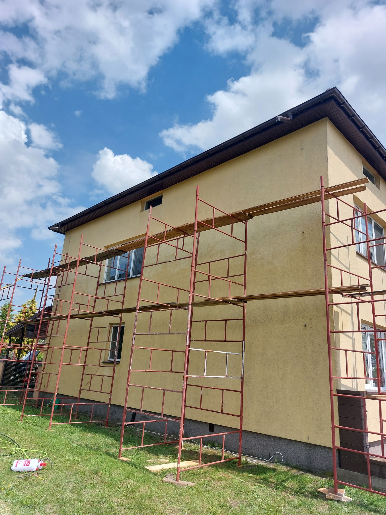 Dwu kondygnacyjny dom w trakcie prac wykończeniowych elewacji, otoczony rusztowaniem z czerwonych rurek, na trawniku leży biały pojemnik z wężykiem.