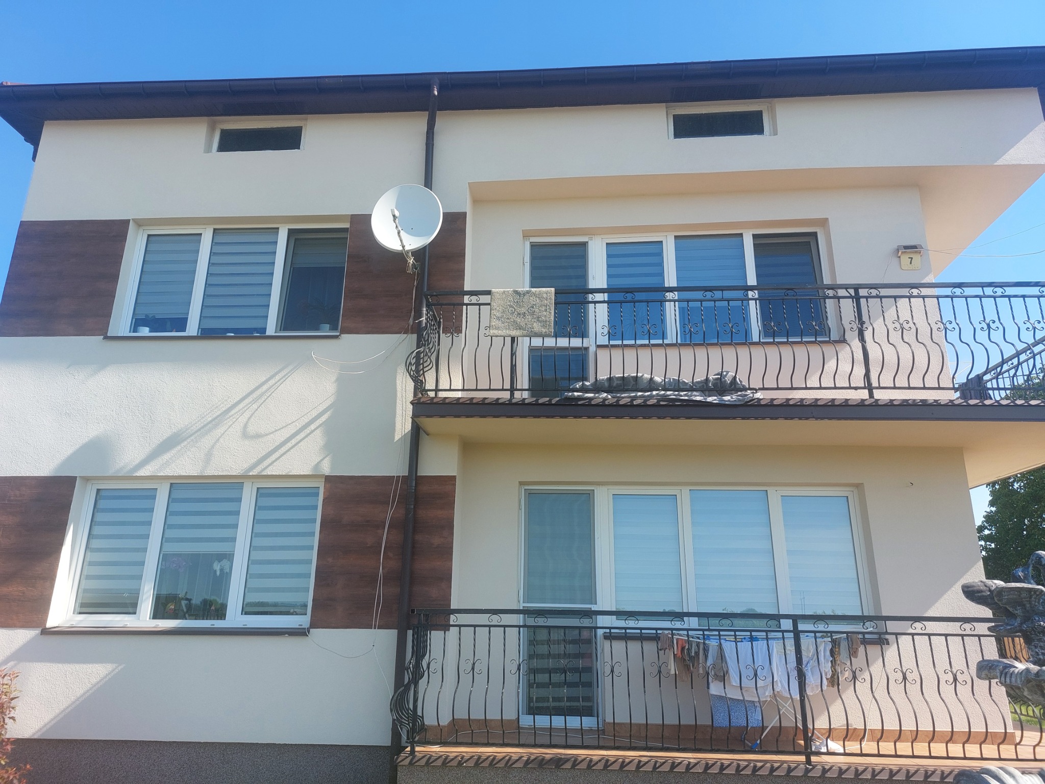 Dwupiętrowy dom z beżową elewacją i brązowymi akcentami, widoczne okna z roletami, balkon z metalową balustradą, antena satelitarna i suszarka z praniem.