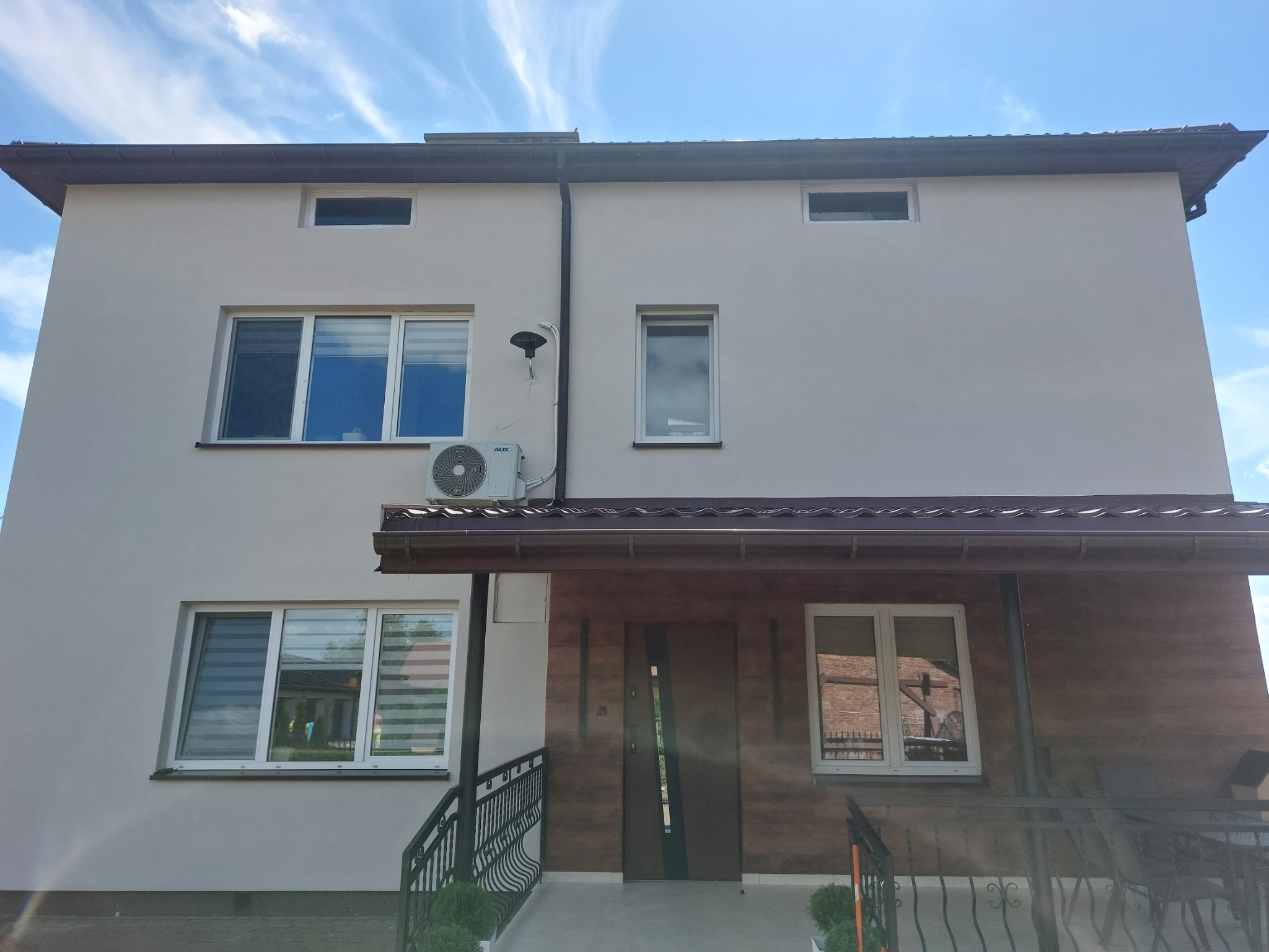 Dwukondygnacyjny dom z jasną elewacją, brązowym dachem i drewnianą wstawką nad wejściem, widoczny klimatyzator na ścianie.