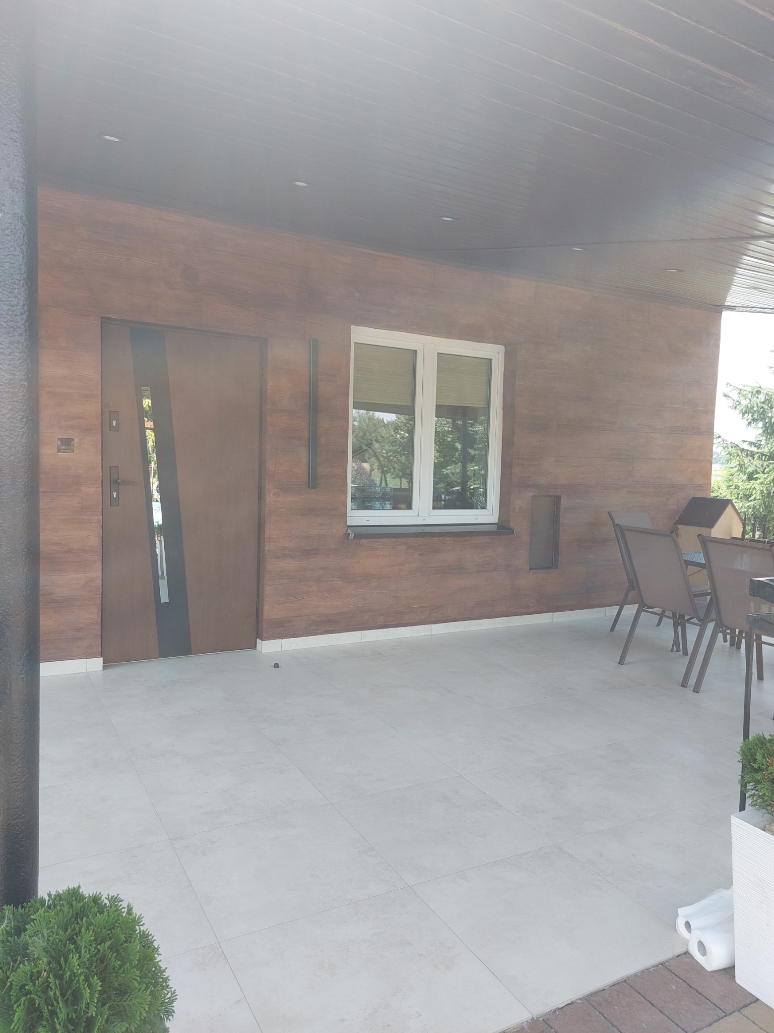 Zadaszony taras z drewnopodobną elewacją, nowoczesnymi drzwiami wejściowymi z przeszkleniem, białymi oknami i jasną, kwadratową terakotą na podłodze, z widocznymi meblami ogrodowymi i zielenią w tle.