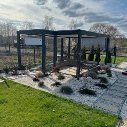 HORTUS - Nowoczesna, grafitowa konstrukcja zadaszenia tarasu, otoczona białym żwirem i zielenią, z widocznym w tle ogrodzeniem i pojazdami dostawczymi.