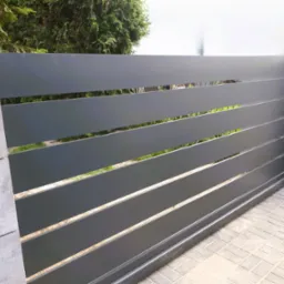 Przesuwna brama wjazdowa z poziomymi panelami w kolorze grafitowym, osadzona na betonowych słupkach, widoczna z poziomu brukowanej nawierzchni.