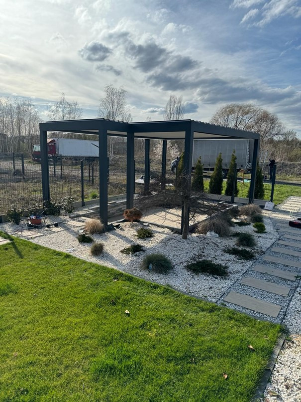 Nowoczesna, grafitowa konstrukcja zadaszenia tarasu, otoczona białym żwirem i zielenią, z widocznym w tle ogrodzeniem i pojazdami dostawczymi.