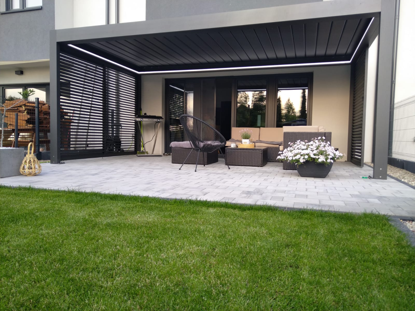 Nowoczesna pergola z czarną konstrukcją i oświetleniem LED, z meblami ogrodowymi na szarym brukowanym patio, widok na zadbany trawnik