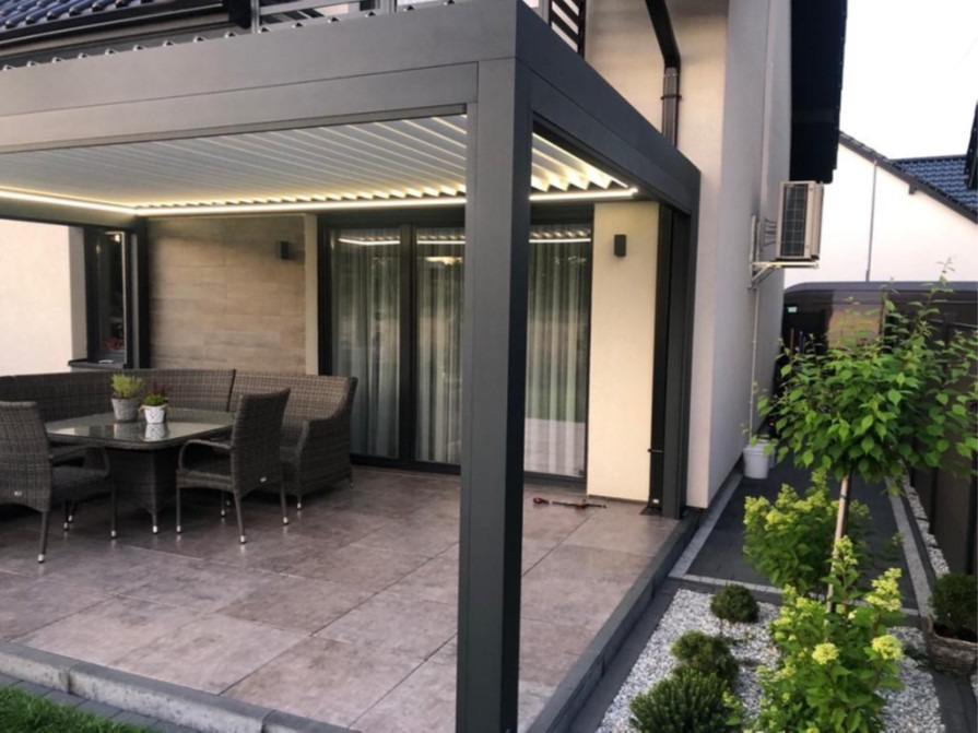 Nowoczesna, szara pergola z oświetleniem LED, zadaszająca taras z meblami ogrodowymi i roślinami ozdobnymi w kamiennym żwirku.