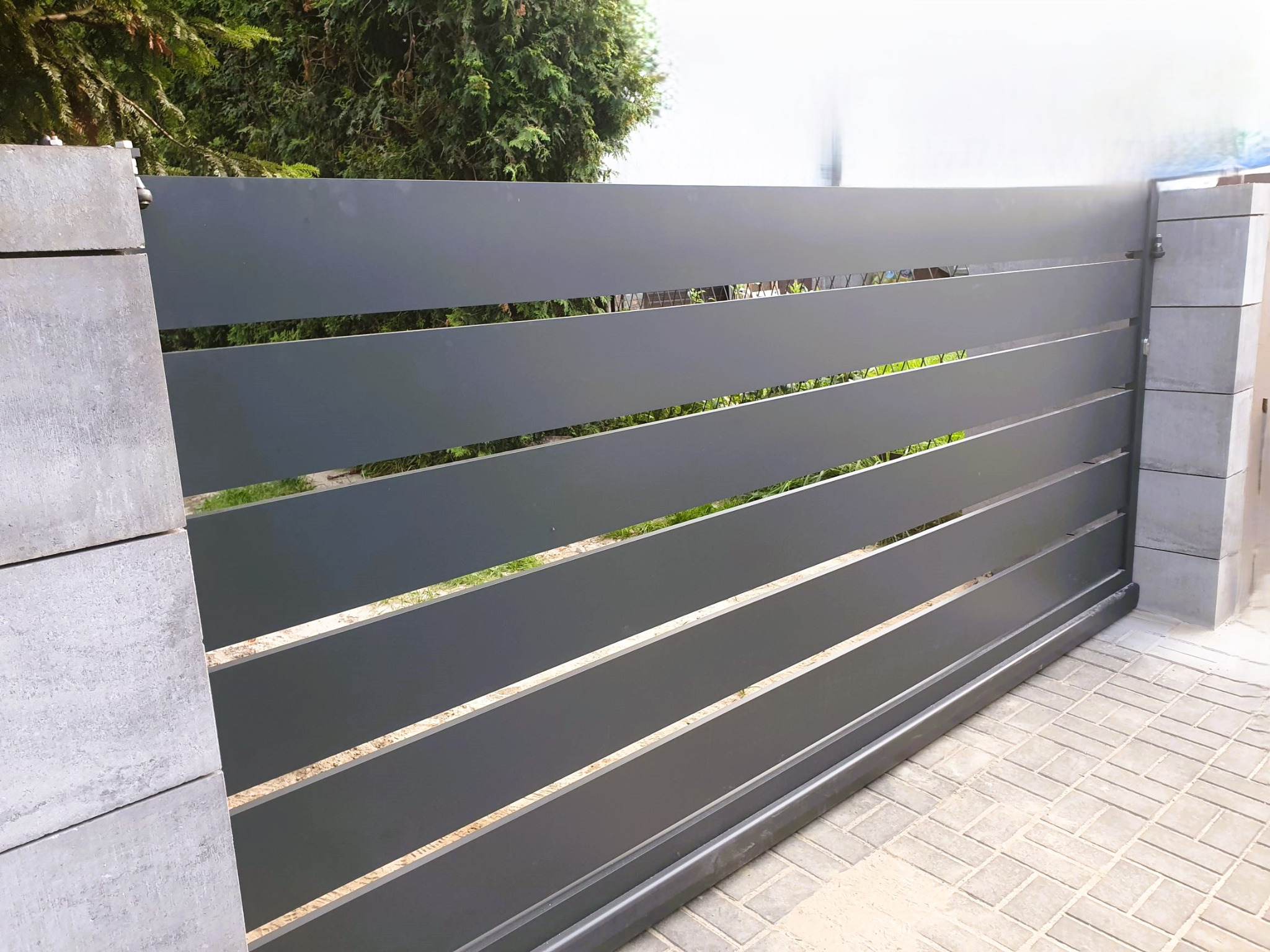 Przesuwna brama wjazdowa z poziomymi panelami w kolorze grafitowym, osadzona na betonowych słupkach, widoczna z poziomu brukowanej nawierzchni.