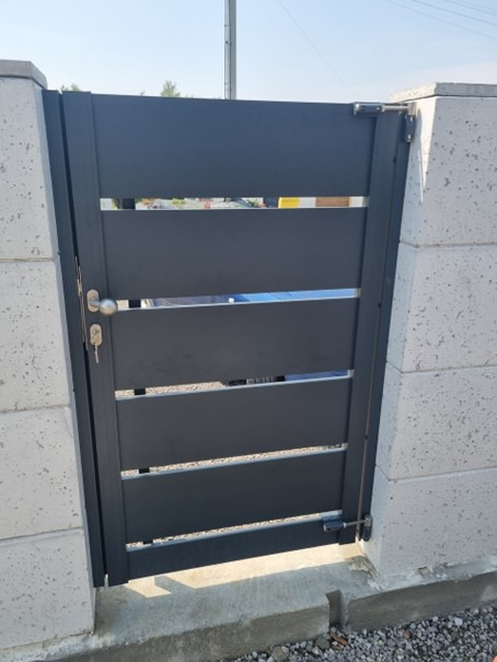 Nowoczesna, grafitowa furtka z poziomymi panelami, zamontowana między betonowymi słupkami, z widocznym zamkiem i kluczem w zamku.