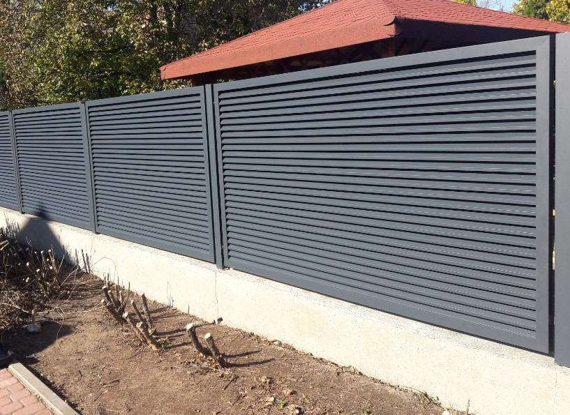 Nowoczesne, szare ogrodzenie z poziomych paneli, widoczne z zewnątrz posesji, z fragmentem altany w tle.