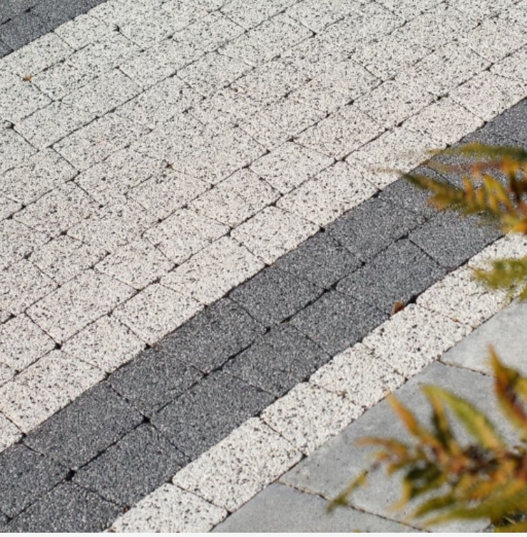Ułożona kostka brukowa w odcieniach szarości i bieli, z fragmentem zieleni w prawym dolnym rogu kadru, prezentująca wzór diagonalny.