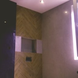 Marcin Wołoszyn - Wnętrze łazienki w trakcie remontu: ściana z płytek ułożonych w jodełkę, wnęka z przygotowanym miejscem na półki, ściana z szarych płytek oraz fragment czarnej ściany z widocznymi otworami...