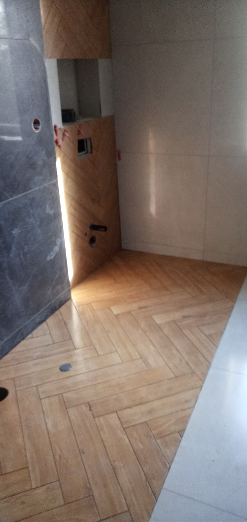 Ułożona w jodełkę podłoga z płytek imitujących drewno w trakcie remontu łazienki, widoczne fragmenty szarej ściany z marmurkowym wzorem i białe płytki na ścianie