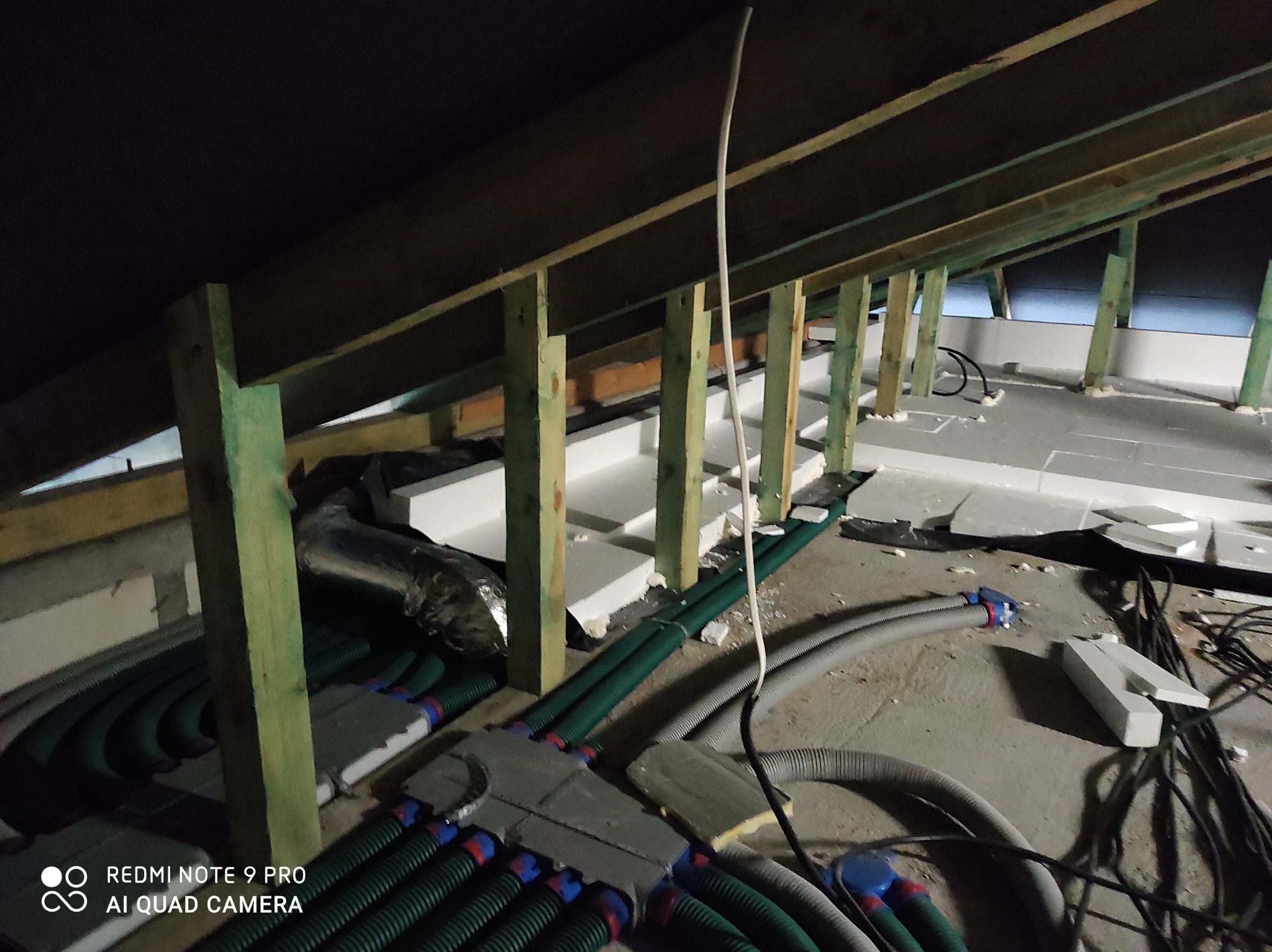 Poddasze w trakcie prac wykończeniowych: widoczne drewniane belki konstrukcyjne, izolacja ze styropianu, liczne przewody wentylacyjne i kable elektryczne ułożone na podłodze.