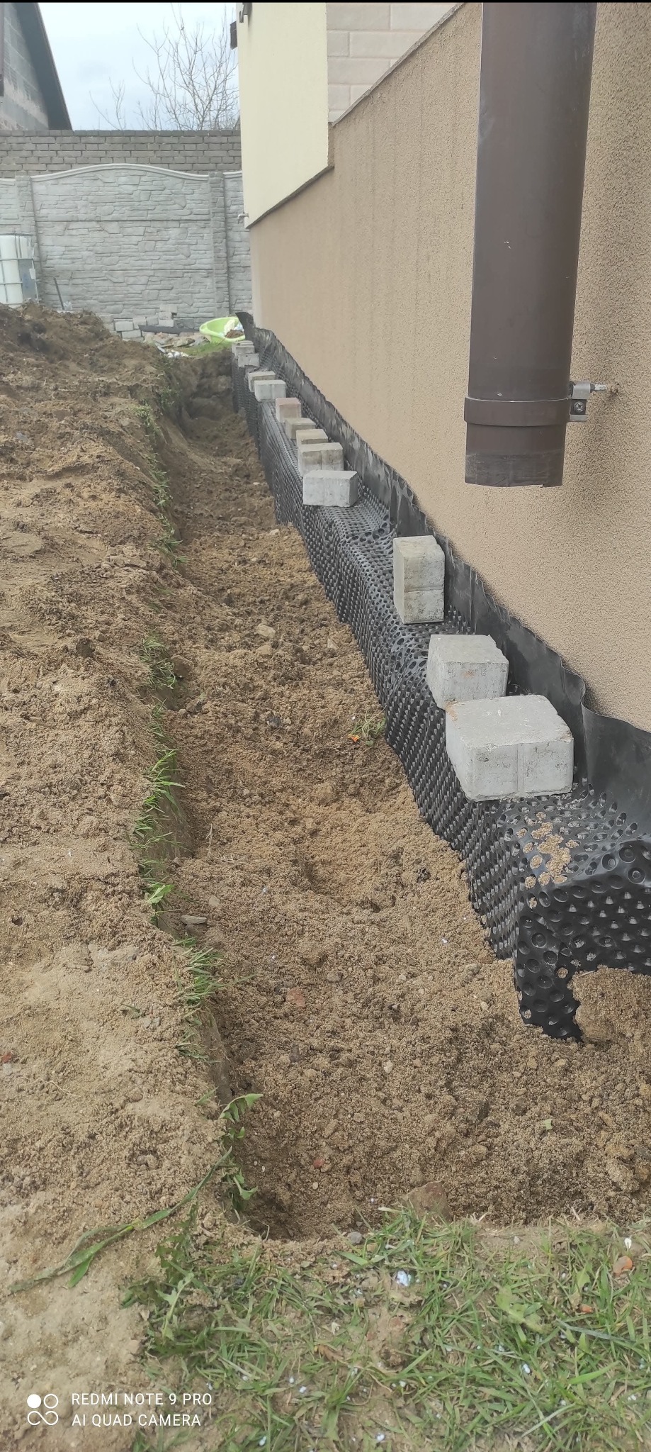 Fundament budynku z izolacją pionową z folii kubełkowej, obłożony betonowymi kostkami, widoczny wykop wzdłuż ściany.