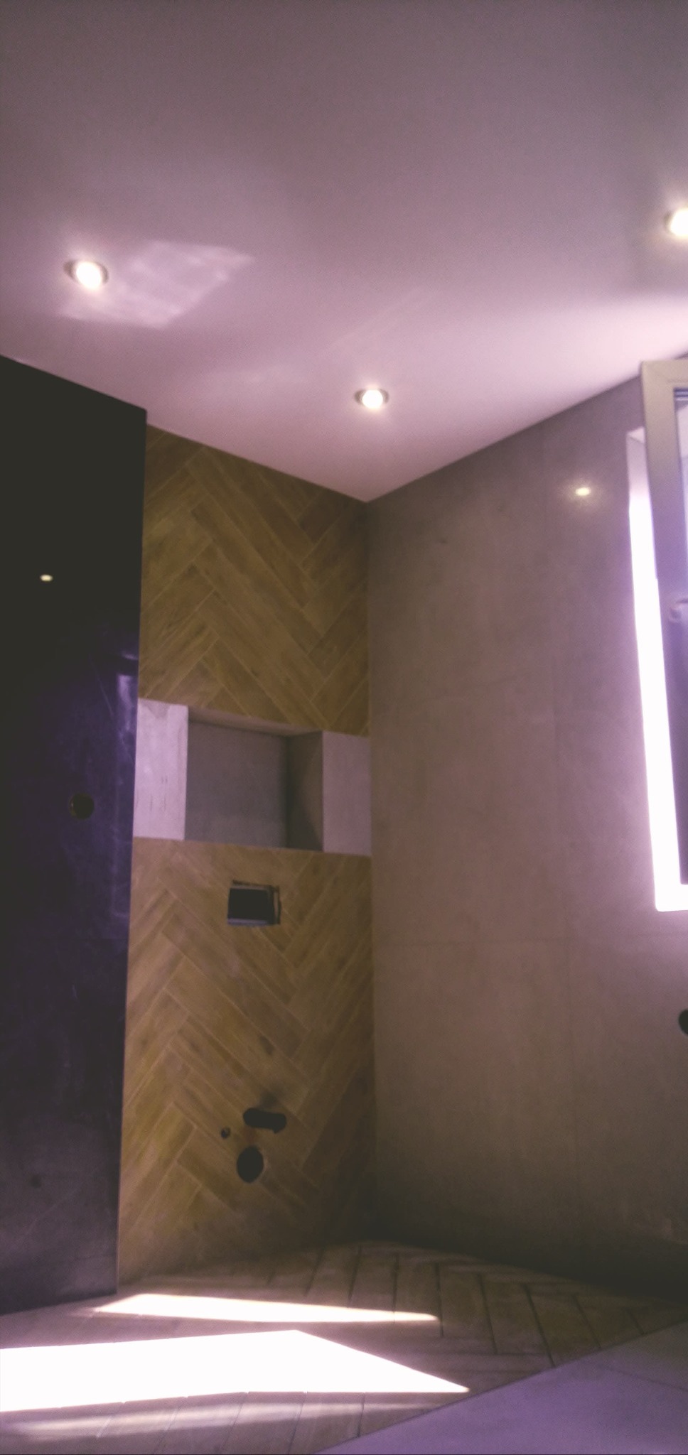 Wnętrze łazienki w trakcie remontu: ściana z płytek ułożonych w jodełkę, wnęka z przygotowanym miejscem na półki, ściana z szarych płytek oraz fragment czarnej ściany z widocznymi otworami...