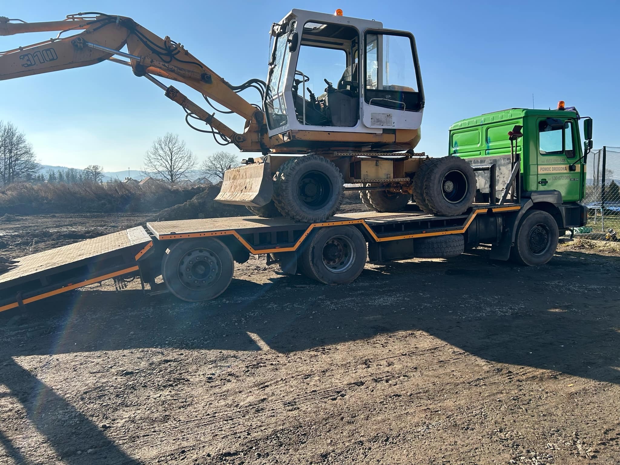 Żółto-biała koparka kołowa marki 310 na niskopodwoziowej platformie transportowej, ciągniętej przez zieloną ciężarówkę pomocy drogowej z widocznymi numerami telefonów na drzwiach, na tle nieba...