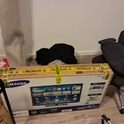 Karton telewizora Samsung LED 40 cali, zabezpieczony taśmą ostrzegawczą, gotowy do transportu podczas przeprowadzki. Widoczne elementy wyposażenia mieszkania.