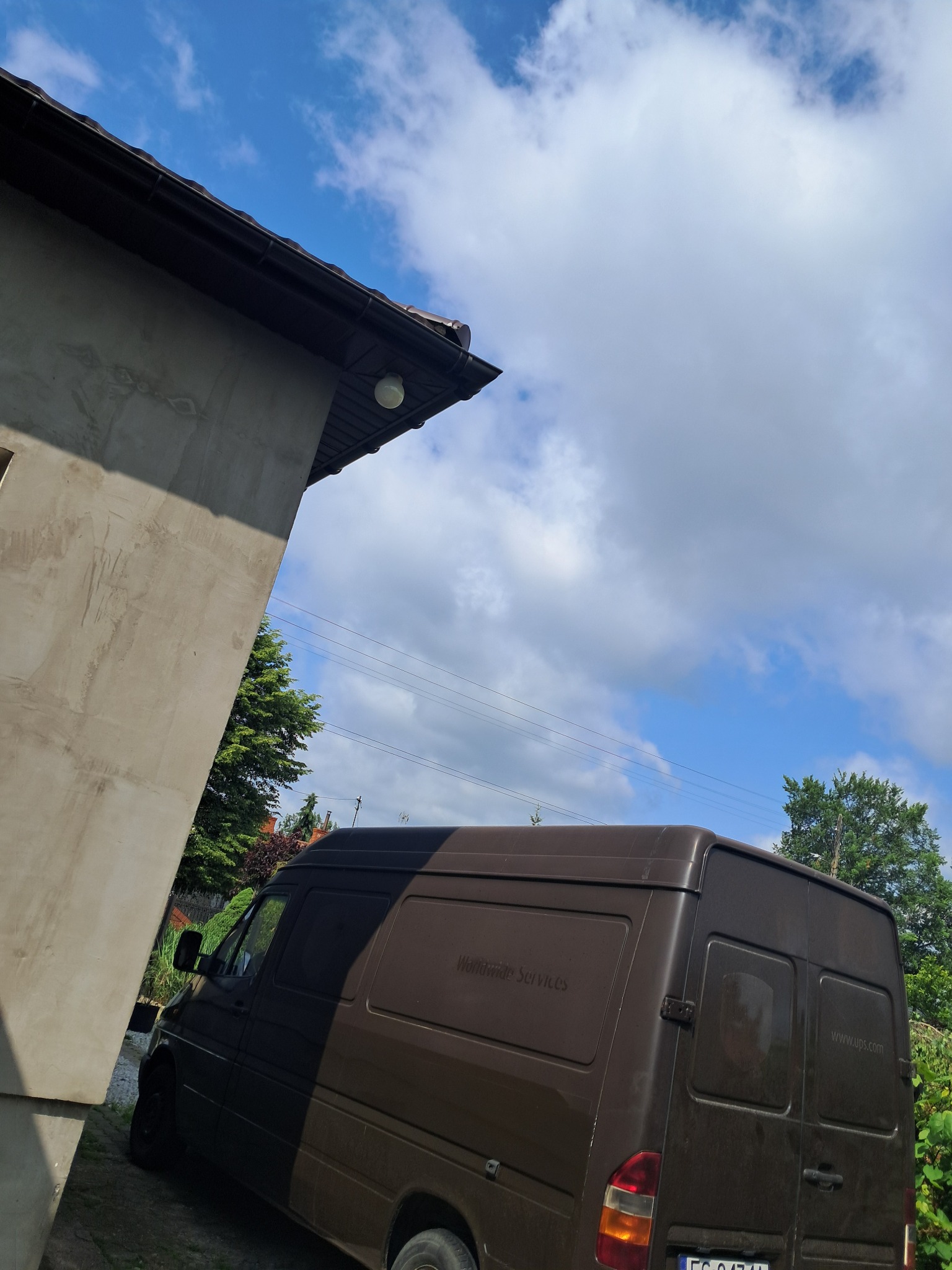 Brązowy bus dostawczy zaparkowany obok budynku z szarą elewacją i widokiem na błękitne niebo z chmurami. Na karoserii widoczny napis 'Worldwide Services'.