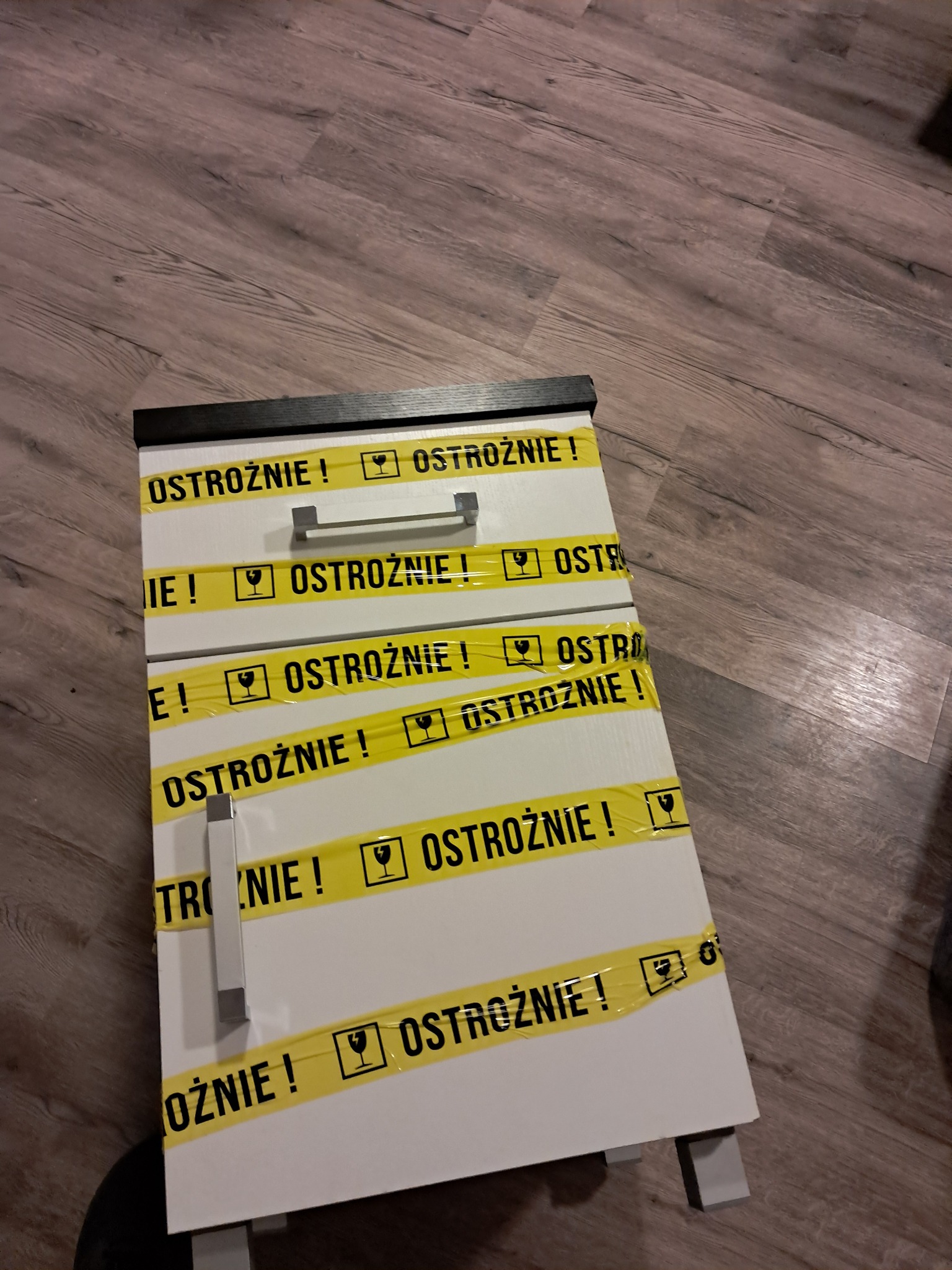 Szafka oklejona taśmą ostrzegawczą z napisem 'Ostrożnie!' i symbolem kieliszka, przygotowana do transportu na podłodze imitującej drewno.
