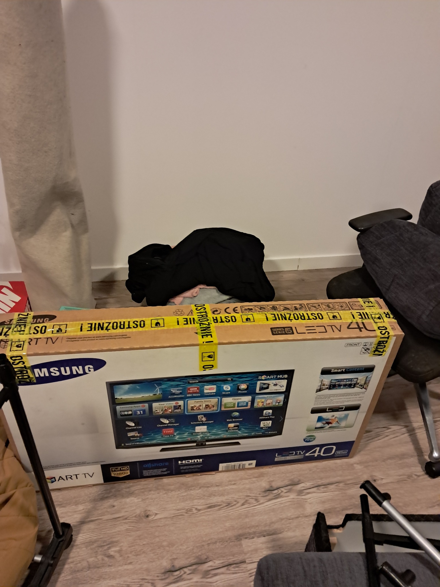 Karton telewizora Samsung LED 40 cali, zabezpieczony taśmą ostrzegawczą, gotowy do transportu podczas przeprowadzki. Widoczne elementy wyposażenia mieszkania.