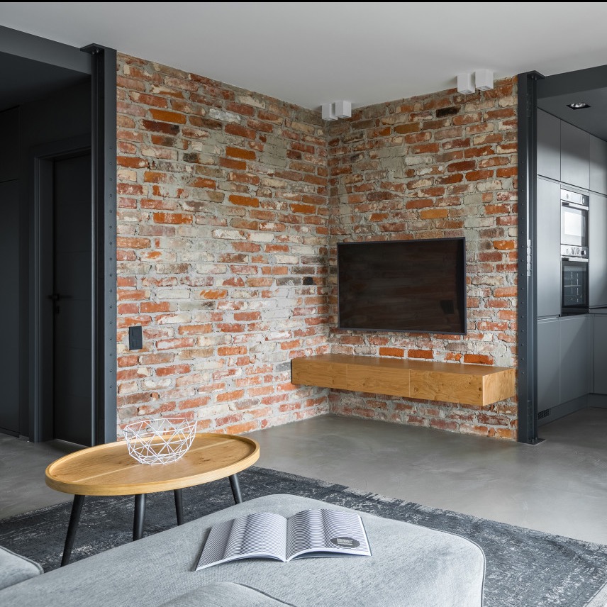 Loftowe wnętrze: ceglana ściana z telewizorem i wiszącą szafką RTV, szara sofa z książką, drewniany stolik kawowy z metalową misą, szara kuchnia w tle.