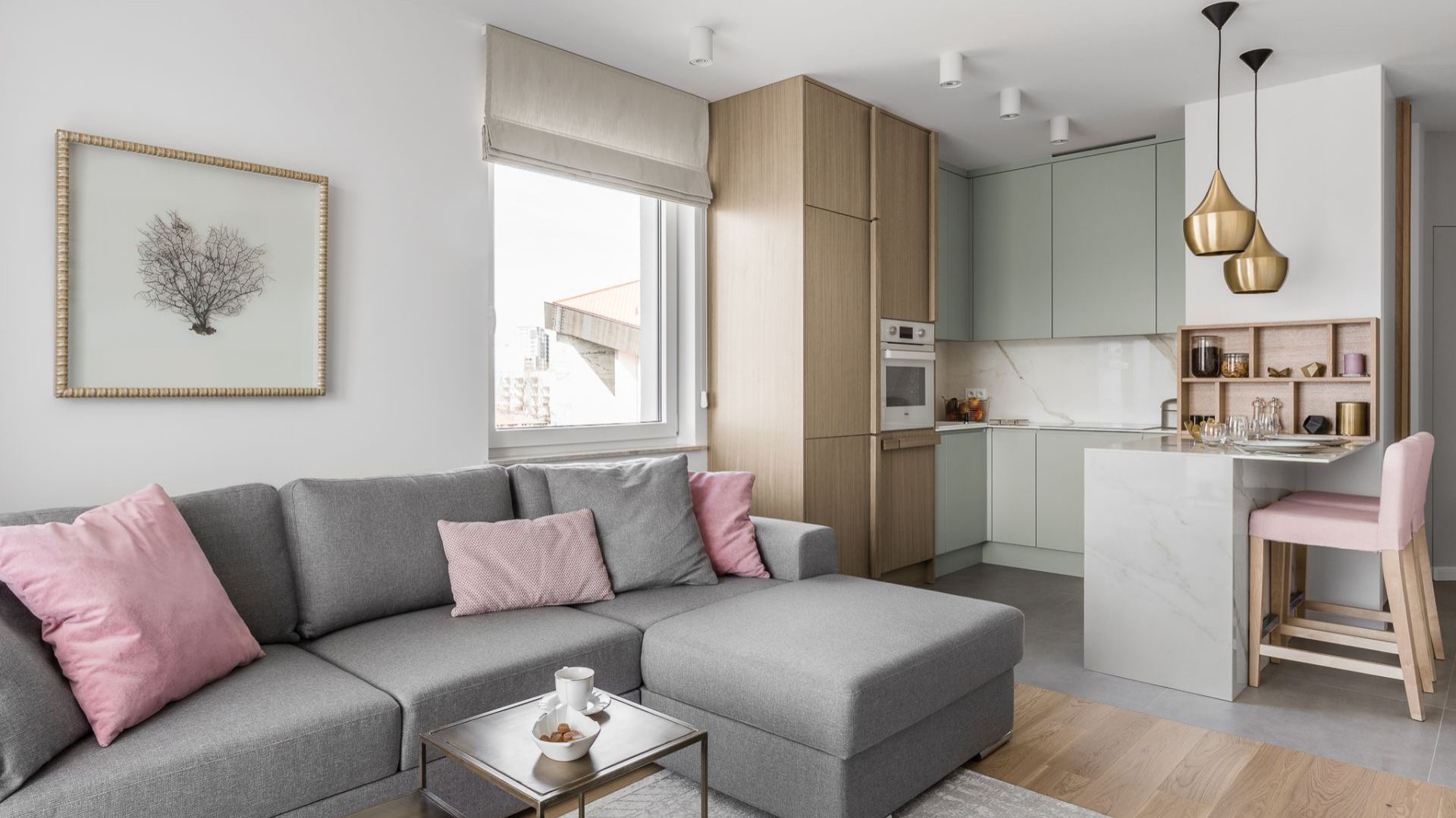 Jasne wnętrze z szarą sofą z różowymi poduszkami, stolikiem kawowym z przekąskami, i kuchnią w odcieniach zieleni i drewna z marmurowym blatem i złotymi lampami wiszącymi.