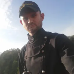 Kominiarz w czarnym uniformie i czapce z napisem 'KOMINIARZ' spogląda w dół, stojąc na dachu z czerwonej dachówki na tle błękitnego nieba i zieleni drzew.