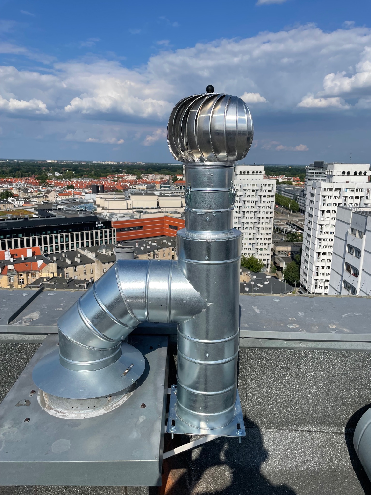 Metalowa turbina wentylacyjna na dachu budynku z widokiem na miasto w słoneczny dzień. Element systemu wentylacji.