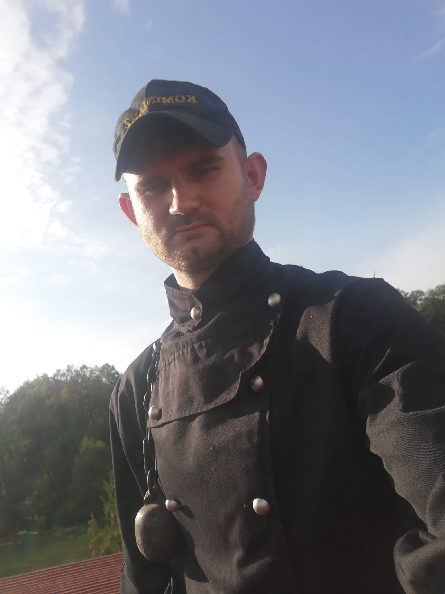 Kominiarz w czarnym uniformie i czapce z napisem 'KOMINIARZ' spogląda w dół, stojąc na dachu z czerwonej dachówki na tle błękitnego nieba i zieleni drzew.