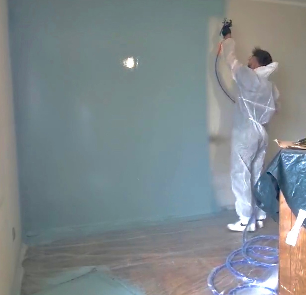 Pracownik w kombinezonie maluje ścianę na niebiesko za pomocą pistoletu natryskowego. Częściowo pomalowana ściana, zabezpieczona folią podłoga i przewód urządzenia.