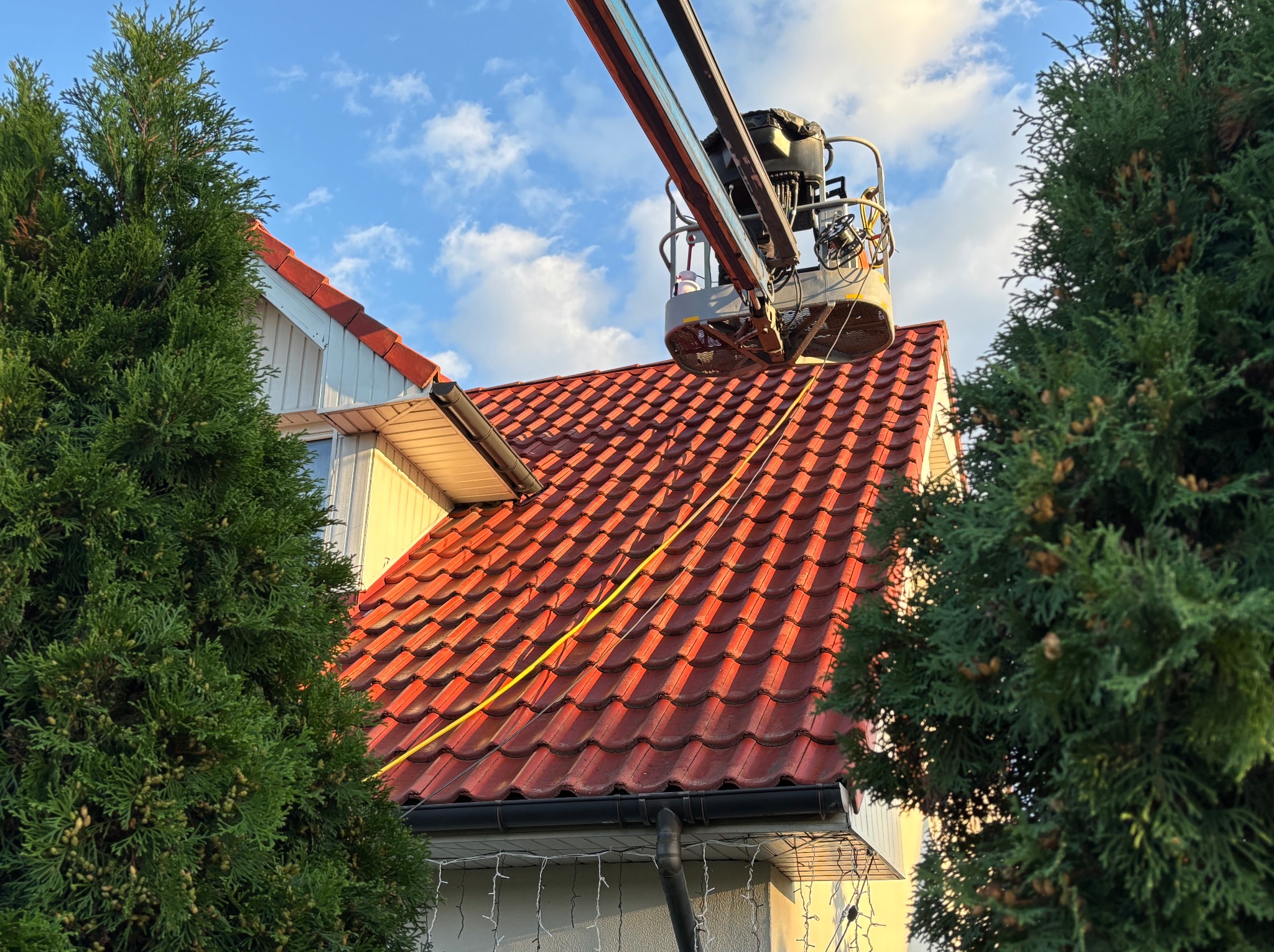 Prace na wysokości: podnośnik koszowy przy czerwonym dachu domu, widoczne narzędzia i wiadro. Przewód zwisa z dachu. Dwa drzewa po bokach kadru.