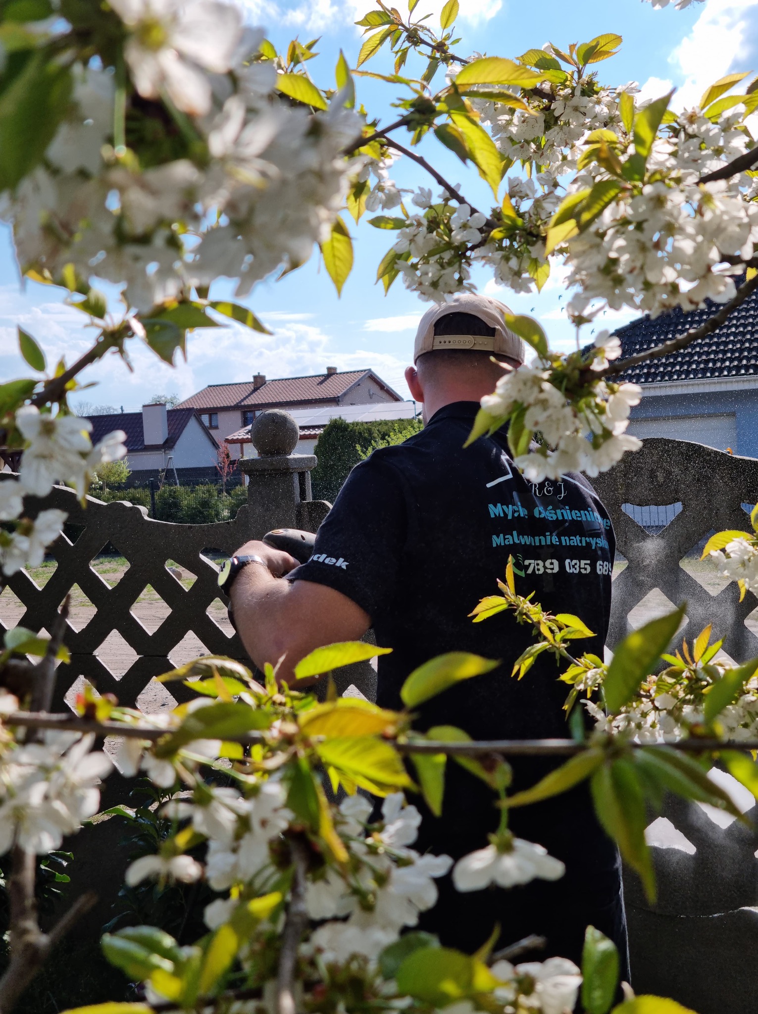 Mężczyzna w czarnej koszulce z logo firmy maluje natryskowo betonowe ogrodzenie przez kwitnące gałęzie drzewa owocowego.