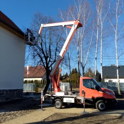 GHY SPÓŁKA Z OGRANICZONĄ ODPOWIEDZIALNOŚCIĄ - Pomarańczowy podnośnik koszowy marki Iveco, z wysięgnikiem ustawionym przy białej ścianie budynku z czerwonym dachem, na tle błękitnego nieba i rzędu wysokich brzóz.