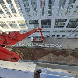 GHY SPÓŁKA Z OGRANICZONĄ ODPOWIEDZIALNOŚCIĄ - Widok z góry na czerwoną platformę podnośnika koszowego przed nowoczesnym budynkiem biurowym z szklaną fasadą podczas prac na wysokości.