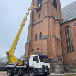 GHY SPÓŁKA Z OGRANICZONĄ ODPOWIEDZIALNOŚCIĄ - Żółty podnośnik koszowy MAN na tle ceglanej wieży kościelnej, platforma z pracownikiem na wysokości okien wieży, pochmurne niebo.