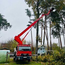 GHY SPÓŁKA Z OGRANICZONĄ ODPOWIEDZIALNOŚCIĄ - Czerwony podnośnik koszowy MAN z wysięgnikiem unosi platformę roboczą w koronie wysokiego drzewa, obok porozrzucane gałęzie, w tle domek dla dzieci