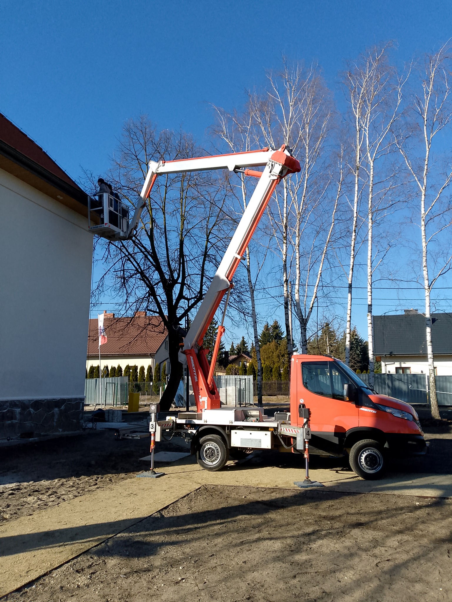 Pomarańczowy podnośnik koszowy marki Iveco, z wysięgnikiem ustawionym przy białej ścianie budynku z czerwonym dachem, na tle błękitnego nieba i rzędu wysokich brzóz.