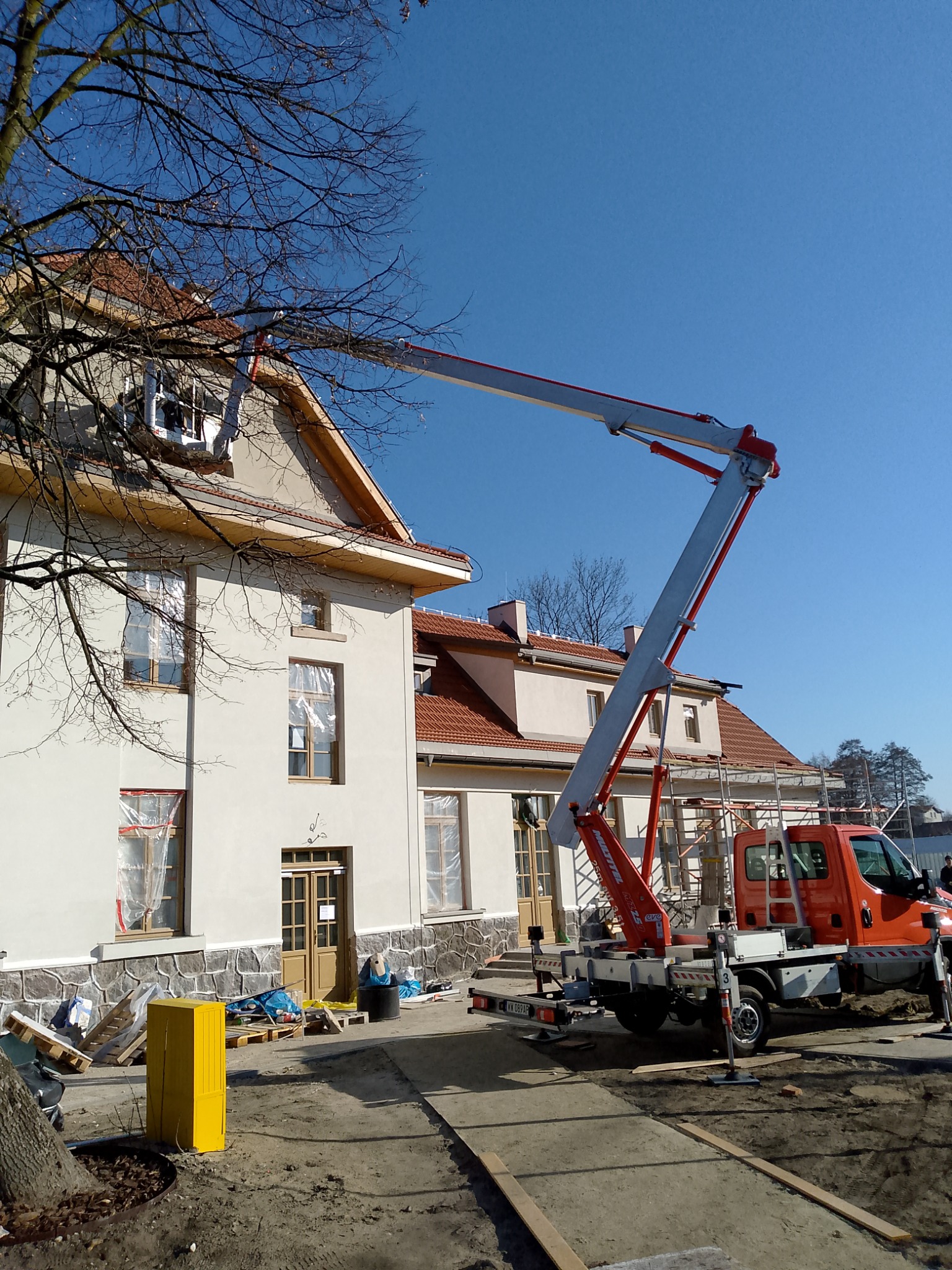 Pomarańczowy podnośnik koszowy Mittal na tle budynku z oknami zabezpieczonymi folią, przygotowanego do prac elewacyjnych w słoneczny dzień.