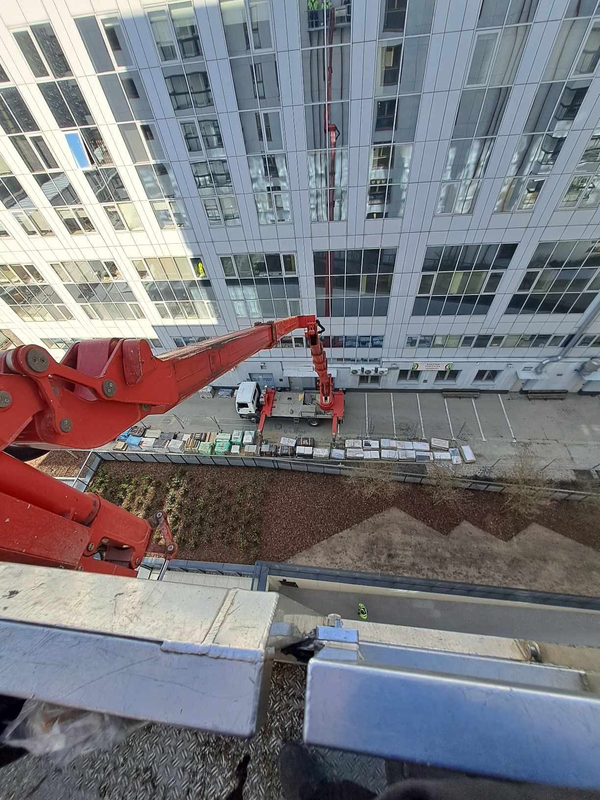 Widok z góry na czerwoną platformę podnośnika koszowego przed nowoczesnym budynkiem biurowym z szklaną fasadą podczas prac na wysokości.