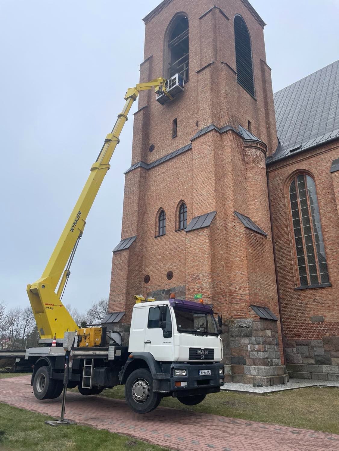 Żółty podnośnik koszowy MAN na tle ceglanej wieży kościelnej, platforma z pracownikiem na wysokości okien wieży, pochmurne niebo.